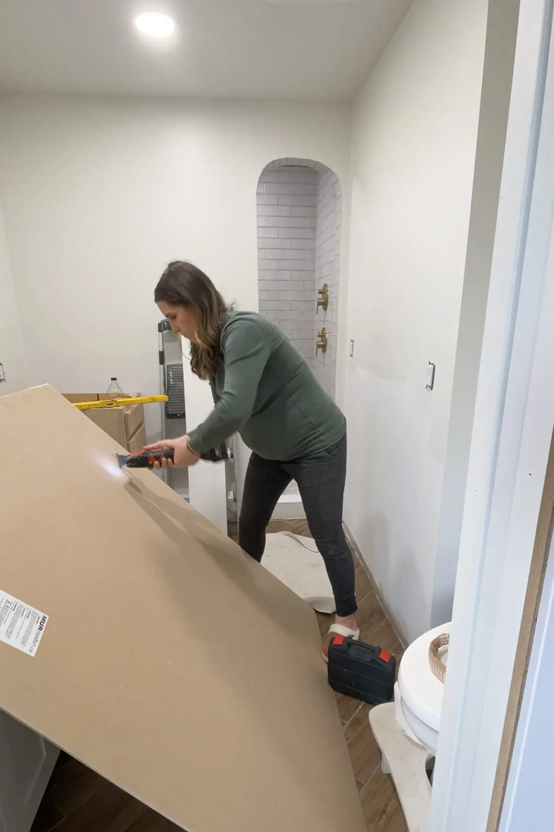 A woman assembling a DIY bathroom renovation, using a power drill to attach a panel to the wall. The unfinished space features a modern arched shower with white subway tiles and brass fixtures. A ladder and construction tools are scattered around, showing the progress of the makeover.
